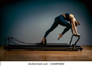 Young girl doing pilates exercises with a reformer bed. Beautiful slim fitness trainer on a reformer gray background, low key, art light, copy space advertising banner