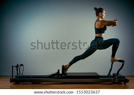 Young girl doing pilates exercises with a reformer bed. Beautiful slim fitness trainer on a reformer gray background, low key, art light, copy space advertising banner