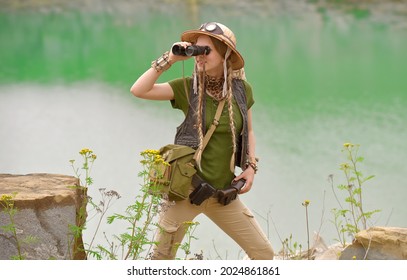 A young girl is dressed up as an explorer. She 
is seen in a green lake environment observing 
the area with her binoculars.