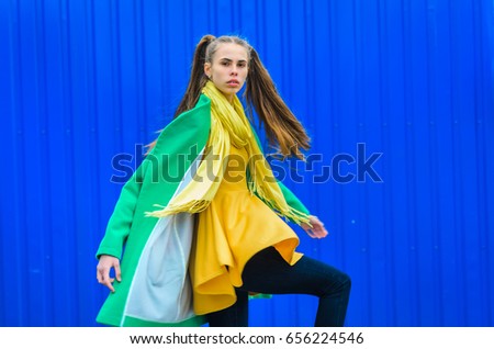 a young girl dressed in a green coat and yellow dress. Pure colors in the photo. girl fun fooling around . against the background of blue wall to show emotions