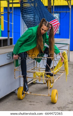 a young girl dressed in a green coat and yellow dress. Pure colors in the photo. girl fun fooling around . the grey shelves, the girl in the yellow truck . is there American flag.