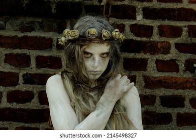 A young girl is dressed as a Halloween horror figure. 
				She wears filthy rags as clothing and has a thorny 
				crown with flowers on her head. She peers into the
				camera with a sad expression on her face. 