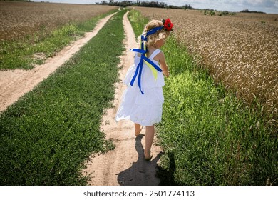 A young girl dressed in a white dress adorned with blue and yellow ribbons joyfully strolls barefoot along a dirt path bordered by fields of golden wheat.  - Powered by Shutterstock