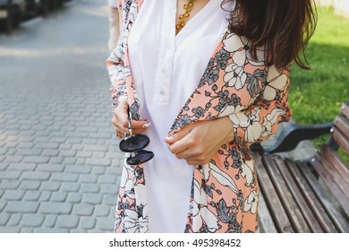 Young Girl In Dress-shirt And Trendy Colorful Cardigan With Dark Glasses In Her Hands. Woman With Long Black Hair In A Pink Cape Walking In The City.