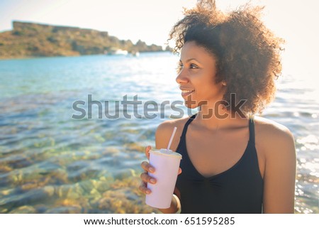 Young girl drinking something fresh. She is at the sea, wearing a black swim suit.