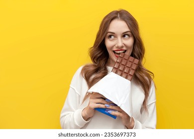 young girl eats and bites big chocolate bar and thinks and dreams on yellow isolated background, happy woman with sweet food looks at copy space