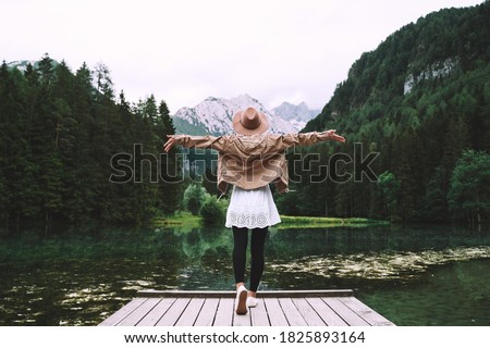 Young girl enjoying beauty of nature looking at mountain lake in Jezersko. Adventure travel in Slovenia, Europe. Woman stands with raised arms on wooden bridge on background with forest and Alps.