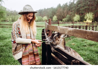 young girl in ethnic dress feeds livestock on a farm. A woman in jeans a woolen sweater and a gray hat feeds a goat on an old ranch. Contact mini zoo in the open air.