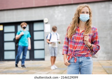 Young Girl In Face Mask With Backpack Standing Near Entrance To School Building. Teenagers Leaving School In Background..