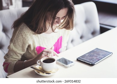 Young girl in a fashionable sweater, sitting in a cafe with a cup of coffee and read messages on the phone and looking pictures on the tablet. - Powered by Shutterstock