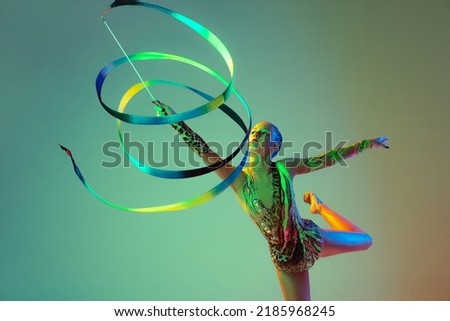 Young girl, female rhythmic gymnast training with multicolored ribbon isolated over green background in neon light. Concept of action, motion, sport, motivation, competition. Copyspace for ad