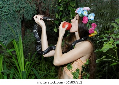 Young girl with floral wreath on her head biting an apple in one hand and holding a serpent in the other