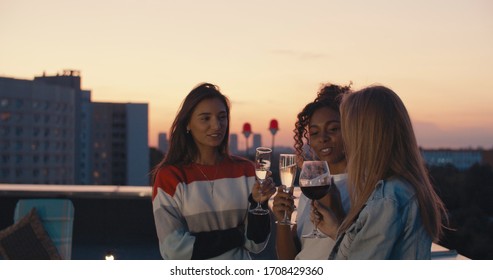 Young Girl Friends Hanging Out With Drinks, Talking, Chatting , Celebrating Birthday. Outdoor Shot Of Young People Toasting Vine At Rooftop Sunset Party.