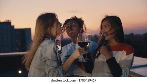 Young Girl Friends Hanging Out With Drinks, Talking, Chatting , Celebrating Birthday. Outdoor Shot Of Young People Toasting Vine At Rooftop Sunset Party.