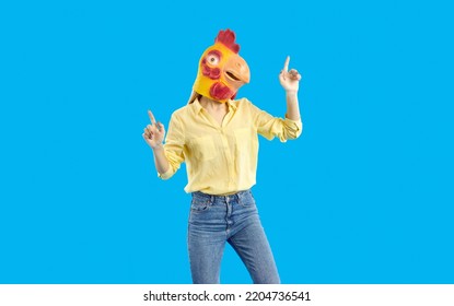 Young girl in funny wacky disguise dancing in the studio. Portrait of crazy woman wearing casual shirt, jeans and goofy yellow chicken, hen or rooster mask dancing isolated on solid blue background