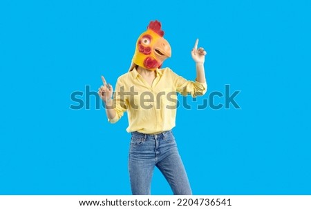 Young girl in funny wacky disguise dancing in the studio. Portrait of crazy woman wearing casual shirt, jeans and goofy yellow chicken, hen or rooster mask dancing isolated on solid blue background