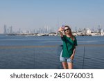 a young girl in a green T-shirt and shorts takes a selfie against the backdrop of the Dubai cityscape