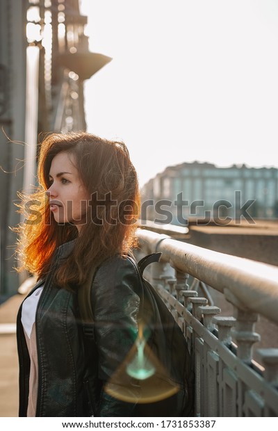 A young girl with hair flying in the wind in a\
leather jacket and a backpack walks on a metal bridge over the Neva\
river in St. Petersburg against the background of a highway in a\
Sunny beautiful sunset