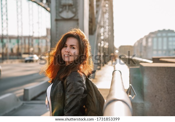 A young girl with hair flying in the wind in a\
leather jacket and a backpack walks on a metal bridge over the Neva\
river in St. Petersburg against the background of a highway in a\
Sunny beautiful sunset