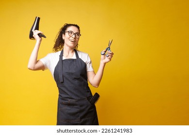 young girl hairdresser in uniform holds a hair dryer and scissors and dances on a yellow background, a woman stylist in an apron shows equipment for work - Powered by Shutterstock