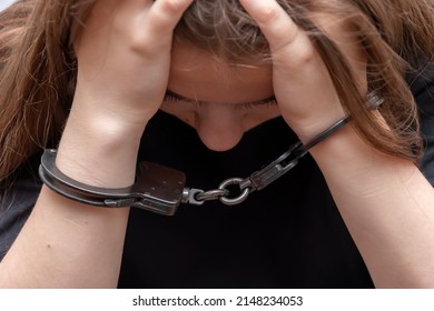 A young girl handcuffed hides her face on a gray background, close-up. Juvenile delinquent in a black T-shirt, criminal liability of minors. - Shutterstock ID 2148234053