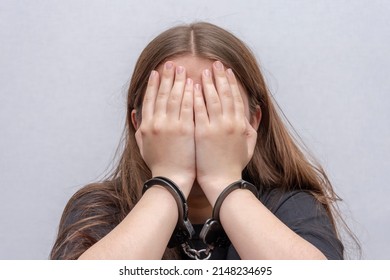 A Young Girl Handcuffed Hides Her Face On A Gray Background, Close-up. Juvenile Delinquent In A Black T-shirt, Criminal Liability Of Minors.