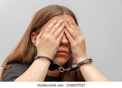A young girl handcuffed hides her face on a gray background, close-up. Juvenile delinquent in a black T-shirt, criminal liability of minors. - Shutterstock ID 2148297037