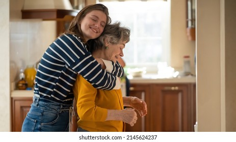A young girl happily hugs her grandmother in a cozy house against the backdrop of the kitchen, her daughter has a good time with her mother, lovingly cares for and supports an elderly relative. - Shutterstock ID 2145624237