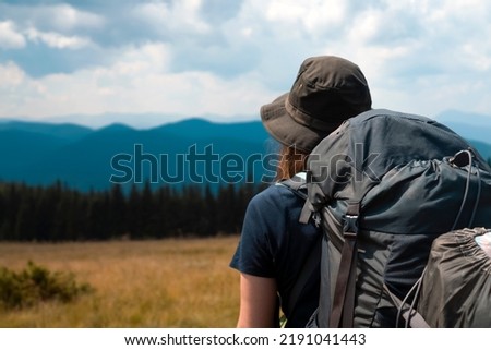 A young girl in a hat and with a backpack goes on an adventure, travels and enjoys nature, a woman looks at a picturesque view with meadows, forest and mountains on a sunny day.