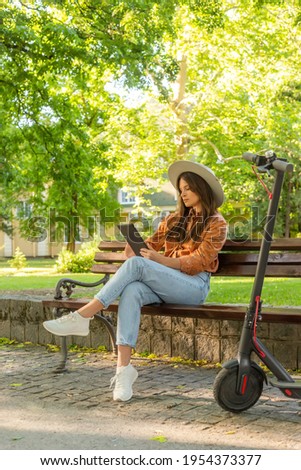 A young girl with a hat is sitting on a park bench and scrolling on the tablet in her hands. An electric scooter is parked next to her, while trees and green colours predominate in the background.