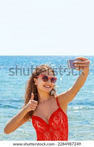 young girl having fun taking smartphone selfie pictures of herself. travel holidays. happy young woman giving hand sign thumbs up on beach.