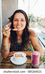 Young Girl Having A Good Morning Healthy Breakfast Smoothie Bowl And A Drink Made Of Super Foods, Fruits, Nuts, Berries, Granola, Shredded Coconut, Acai