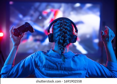 Young girl in headphones plays a video game on the TV in the dark room. Gamer with a joystick. Online gaming with friends, competitions, win. Fun entertainment. Teenagers play puzzles games. Back view - Shutterstock ID 1691902558
