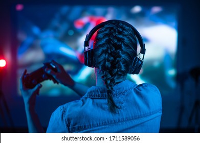 Young girl in headphones plays a video game on a big screen projector or TV. Gamer with a joystick. Online games with friends, competitions, wins. Fun entertainment. Teenagers play arcade. Back view - Shutterstock ID 1711589056