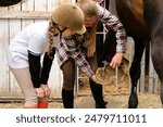 A young girl in a helmet watches an instructor clean a horse