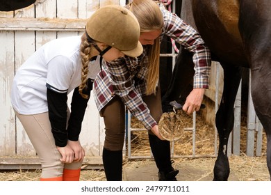 A young girl in a helmet watches an instructor clean a horse's hoof. Routine on a farm, caring for animals in the countryside - Powered by Shutterstock