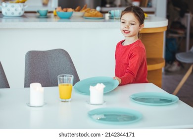 A Young Girl Helps To Set The Family Dining Room Table. Preschooler Kid Helping Mother To Set Table In Kitchen, Carrying The Plates, Kitchen Interior. 