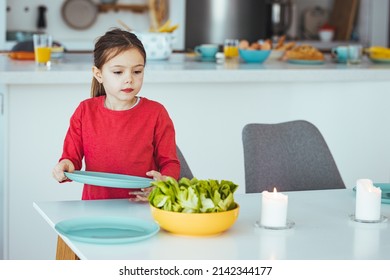 A Young Girl Helps To Set The Family Dining Room Table. Preschooler Kid Helping Mother To Set Table In Kitchen, Carrying The Plates, Kitchen Interior. 