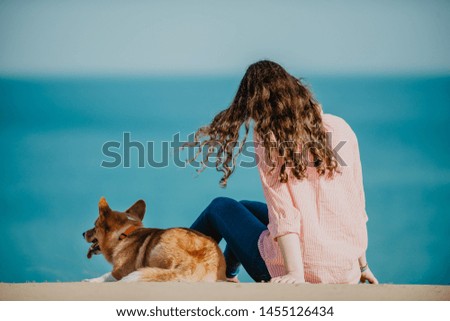 Young girl and her corgi dog sitting on sand looking at the ocean, the girl's hair flying in the wind