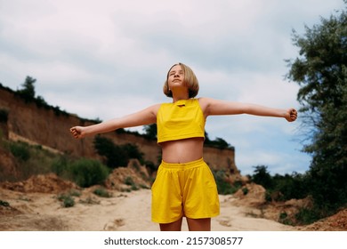 A young girl with her eyes closed, in a yellow summer suit, shorts and a tank top, stands among the sandy hills and spreads her arms to the sides.