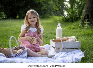 Young girl holding a bread roll, smiling at a picnic setup in a shaded park area, sitting on a blanket. - Powered by Shutterstock