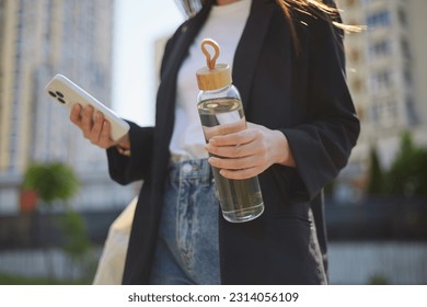 Young girl holding a glass bottle of water and a modern smart phone in hands. Unrecognizable female person walking in the city street with a gadget and a reusable water bottle - Powered by Shutterstock