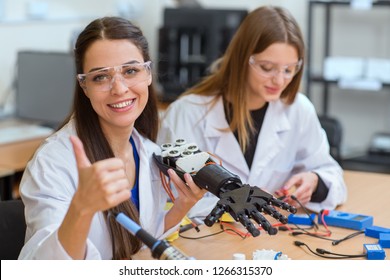Young girl holding plastic hand and gesturing thumb up. Beautiful female scientist, wearing in white uniform, checking invention, smiling and looking at camera while her colleague working behind.