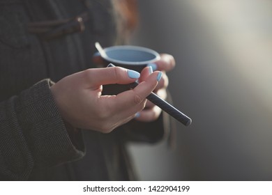 Young Girl Holds Electronic Cigarette In Hand.Woman Smokes Vaping Device And Drinks Cheal Instant Coffee Drink Outdoor.Quit Smoking And Drinking Caffeine.Unhealthy Lifestyle And Bad Nutrition Concept