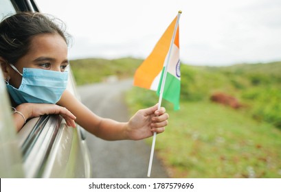 Young girl kid with medical mask holding Indian flag in moving car window - Concept of celebrating Independence or republic day during coronavirus or covid-19 pandemic.
