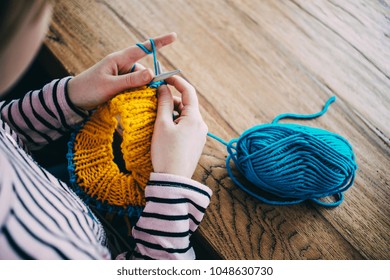 Young Girl Knitting A Circle Scarf With Yellow And Blue Coloured Yarn. Sitting At The Wooden Table, Close Up Of The Knitting Needles.