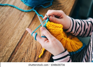 Young girl knitting a circle scarf with yellow and blue coloured yarn. Sitting at the wooden table, close up of the knitting needles.