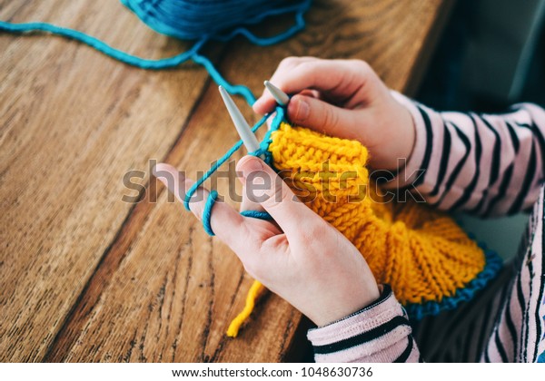 Young girl knitting a circle scarf with yellow\
and blue coloured yarn. Sitting at the wooden table, close up of\
the knitting needles.