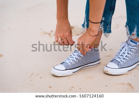 Young girl lacing up tennis shoes on sand beach. Close up of grey sneakers with white shoelaces. Tanned skin, vacation and travel concept