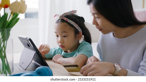 Young Girl Little Kid Help Mom Pay School Charge Tuition Term Fee Or Tax Bill On Web Social Media App Relax Smile At Home Sofa. Asia People Happy Playful Fun Buy Gift Card On Online Shop On Laptop.